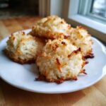 Four golden-brown Vegan Coconut Macaroons piled on a white plate near a window.