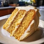 A close-up slice of moist, yellow Vegan Eggnog Cake With Buttercream frosting layers on a light plate.