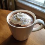 A white mug filled with rich Vegan Hot Cocoa With Coconut Cream, sitting on a wooden table near a window.