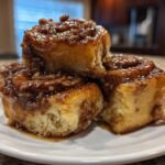 Close-up of gooey Vegan Maple Pecan Sticky Buns stacked on a white plate, dripping with caramel glaze.