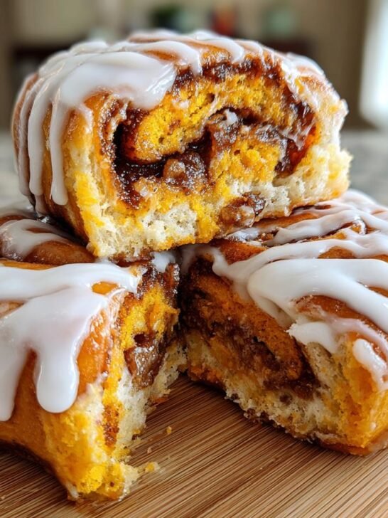 A stack of two Vegan Pumpkin Pie Cinnamon Rolls, one cut open to show the filling, drizzled with thick white icing.