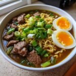 A close-up of a white bowl filled with rich broth, wavy noodles, chunks of beef, and topped with green onions and a soft-boiled egg for amazing Beef Ramen Noodles.