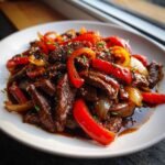 A close-up of glossy, saucy Black Pepper Beef Stir Fry with bright red bell peppers and onions on a white plate.