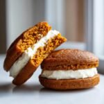Two delicious Pumpkin Whoopie Pies, one whole and one cut in half showing the thick cream cheese filling.