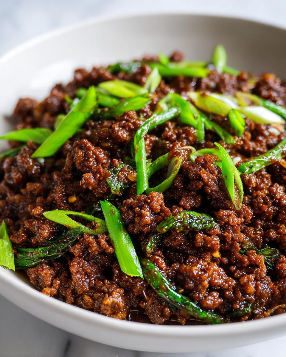 A close-up of rich, dark Asian Ground Beef topped generously with bright green sliced scallions in a white bowl.