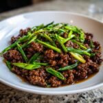 A close-up of saucy Asian ground beef topped generously with bright green, sliced scallions served on a white plate.