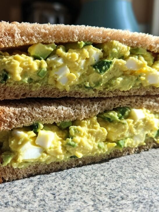 Two halves of an Avocado Egg Salad Sandwich stacked, showing creamy filling with chunks of egg and avocado on whole wheat bread.