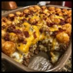 A scoop has been taken from a hot Bacon Cheeseburger Tater Tot Casserole showing melted cheese and ground beef.