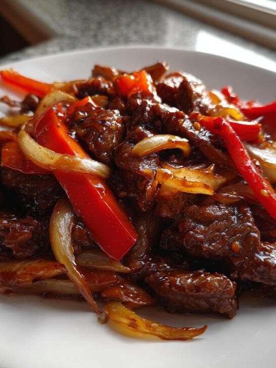A close-up of a serving of Amazing Black Pepper Beef Stir Fry with glossy sauce, red peppers, and onions on a white plate.