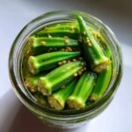 Overhead shot of bright green pickled okra spears packed tightly in a glass jar with pickling brine and mustard seeds.