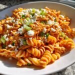 A close-up of Buffalo Chicken Pasta Salad featuring rotini pasta, shredded chicken in orange sauce, topped with feta cheese and green onions.