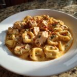A close-up of Cajun Chicken Tortellini With Creamy Garlic Sauce in a white bowl, topped with red pepper flakes.