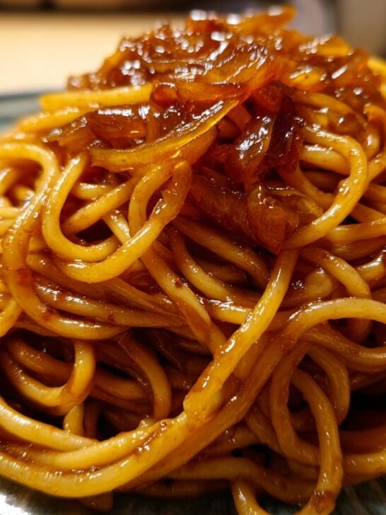 A close-up shot of a mound of glossy spaghetti tossed in sauce, topped with rich, dark caramelized onion pasta.