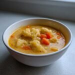 A white bowl filled with creamy, orange-hued Cauliflower Cheddar Vegetable Soup featuring large chunks of cauliflower and bright orange carrots.