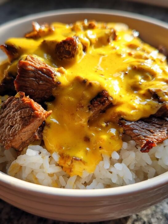 A white bowl filled with white rice, topped with chunks of seasoned steak and smothered in bright yellow queso sauce, creating a Cheesy Steak Queso Rice Bowl.