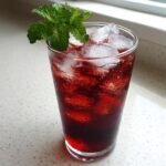 A tall glass filled with vibrant red Cherry Iced Tea, loaded with ice cubes and garnished with fresh mint.
