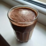 A close-up of a rich, dark Chocolate Avocado Smoothie with foam on top, sitting on a white windowsill.