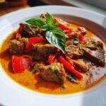 Close-up of tender beef chunks in a vibrant orange Coconut Red Curry Drip Beef sauce, garnished with red peppers and fresh basil.