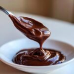 A spoonful of thick, glossy Homemade Nutella Recipe dripping back into a white bowl.
