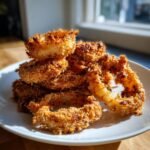 A close-up stack of golden brown, crispy homemade onion rings piled high on a white plate.