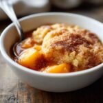 Close-up of a serving of warm Crock Pot Peach Cobbler in a white bowl with a spoon.