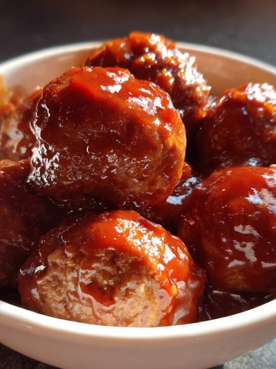 Close-up of several Crockpot Hawaiian Meatballs coated in a thick, glossy, reddish-brown sauce, served in a small white bowl.