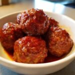 Close-up of several glossy Crockpot Hawaiian Meatballs coated in a thick, sweet and savory red sauce in a white bowl.