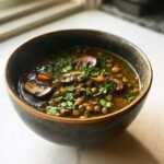A close-up of a dark bowl filled with rich French Lentil And Mushroom Soup, garnished with fresh parsley and sliced mushrooms.
