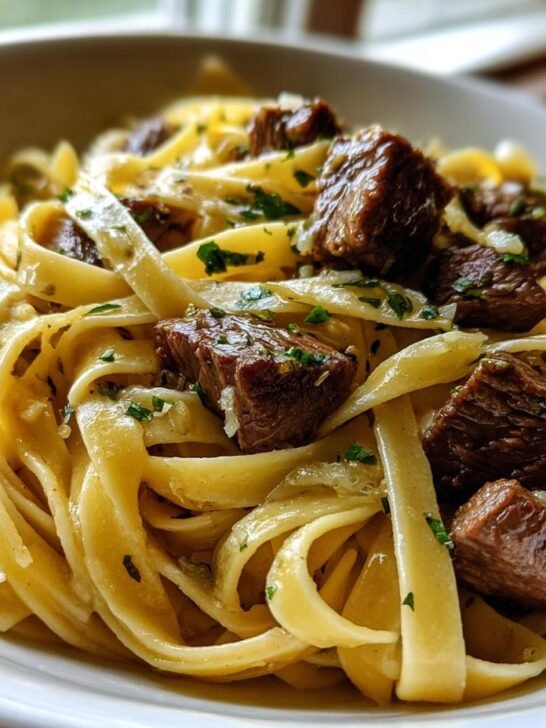 Close-up of a white bowl filled with fettuccine pasta tossed in a light sauce with chunks of seared beef, representing the Garlic Steak Pasta.