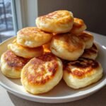 A tall stack of fluffy, golden brown Pancake Bites piled high on a white plate near a window.