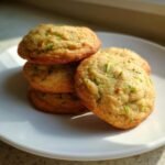 A stack of four golden brown Zucchini Cookies resting on a white plate, showing visible green shreds of zucchini throughout.