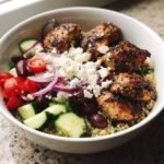 Close-up of a vibrant Greek Chicken Quinoa Bowl topped with grilled chicken, feta, cucumbers, tomatoes, olives, and red onion.