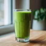 A tall glass filled with a vibrant Green Avocado Smoothie sitting on a wooden table near a window.