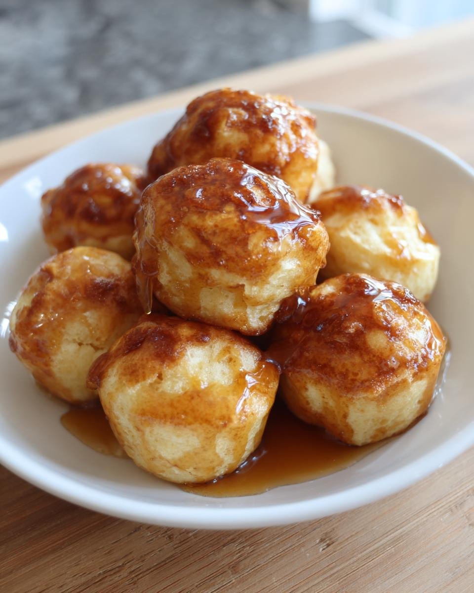 A close-up of several golden Honey Butter Cornbread Poppers stacked in a white bowl and drizzled with honey butter glaze.
