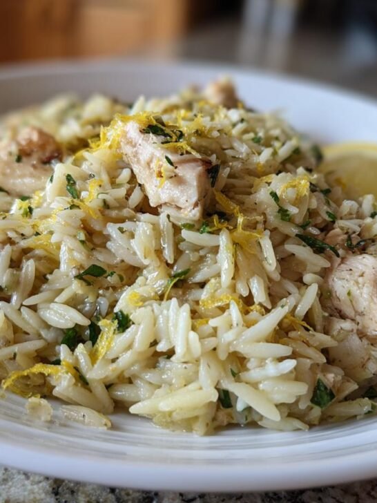 Close-up of a white bowl filled with Lemon Feta Chicken And Orzo, garnished with lemon zest and a lemon wedge.