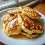 A stack of golden brown Lemon Ricotta Pancakes drizzled generously with syrup on a white plate.