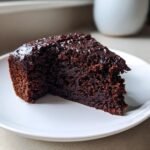 A close-up of a moist, dark slice of Chocolate Zucchini Cake topped with rich, glossy chocolate frosting on a white plate.