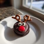 A close-up of an Oreo Reindeer Cookie decorated with chocolate, candy eyes, a red nose, and pretzel antlers.