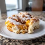 A close-up of a single, gooey Peach Cobbler Cinnamon Roll topped with white icing, sitting on a white plate.