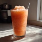 A tall glass filled with vibrant orange Peach Lemonade Slush, topped with crushed ice and a straw.