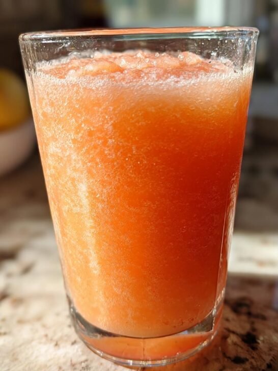 A tall glass filled with bright orange Peach Lemonade Slush, resting on a granite countertop.