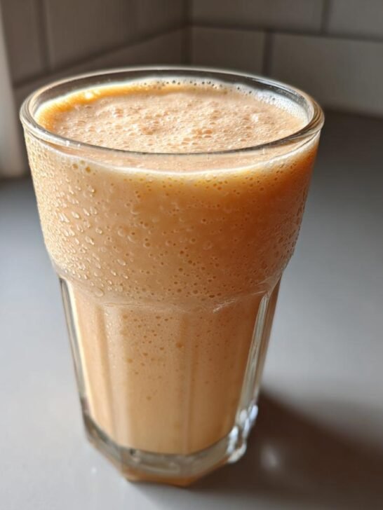 A tall glass filled with a creamy, orange-hued Peach Yogurt Smoothie sitting on a light countertop.