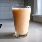 A tall glass filled with a thick, frosty Peach Yogurt Smoothie sitting on a light countertop.