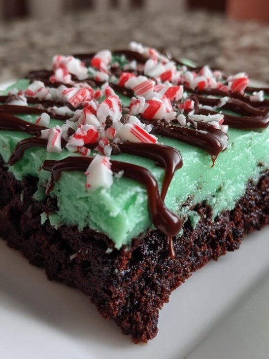 A square of rich chocolate brownie topped with bright green mint frosting, chocolate drizzle, and crushed candy canes for Peppermint Hot Chocolate Brownies.