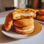 A stack of three soft Pumpkin Cheesecake Cookies, with the top one cut in half showing the thick white cream cheese filling.