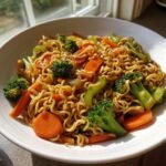 A white bowl filled with savory Ramen Noodle Stir Fry featuring wavy noodles, bright broccoli florets, and sliced carrots.
