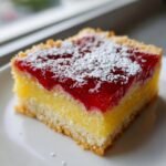 Close-up of a square slice of Raspberry Lemon Bars showing a crust, bright yellow lemon filling, and vibrant red raspberry topping dusted with powdered sugar.