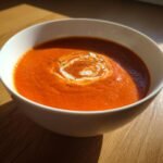 A close-up of vibrant orange Roasted Tomato Basil Soup, garnished with a swirl of cream and black pepper.