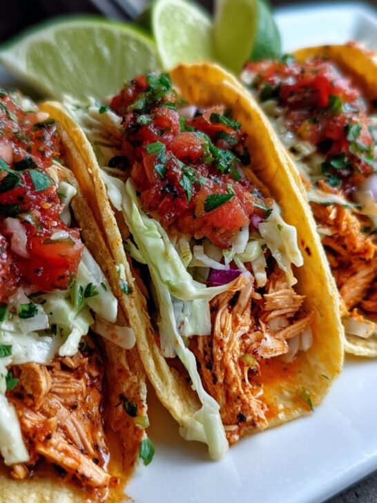 Four delicious Salmon Tacos lined up on a white plate, topped with slaw and fresh salsa, with lime wedges in the background.