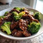 A close-up shot of glossy, saucy Sesame Beef And Broccoli topped with white sesame seeds in a white bowl.